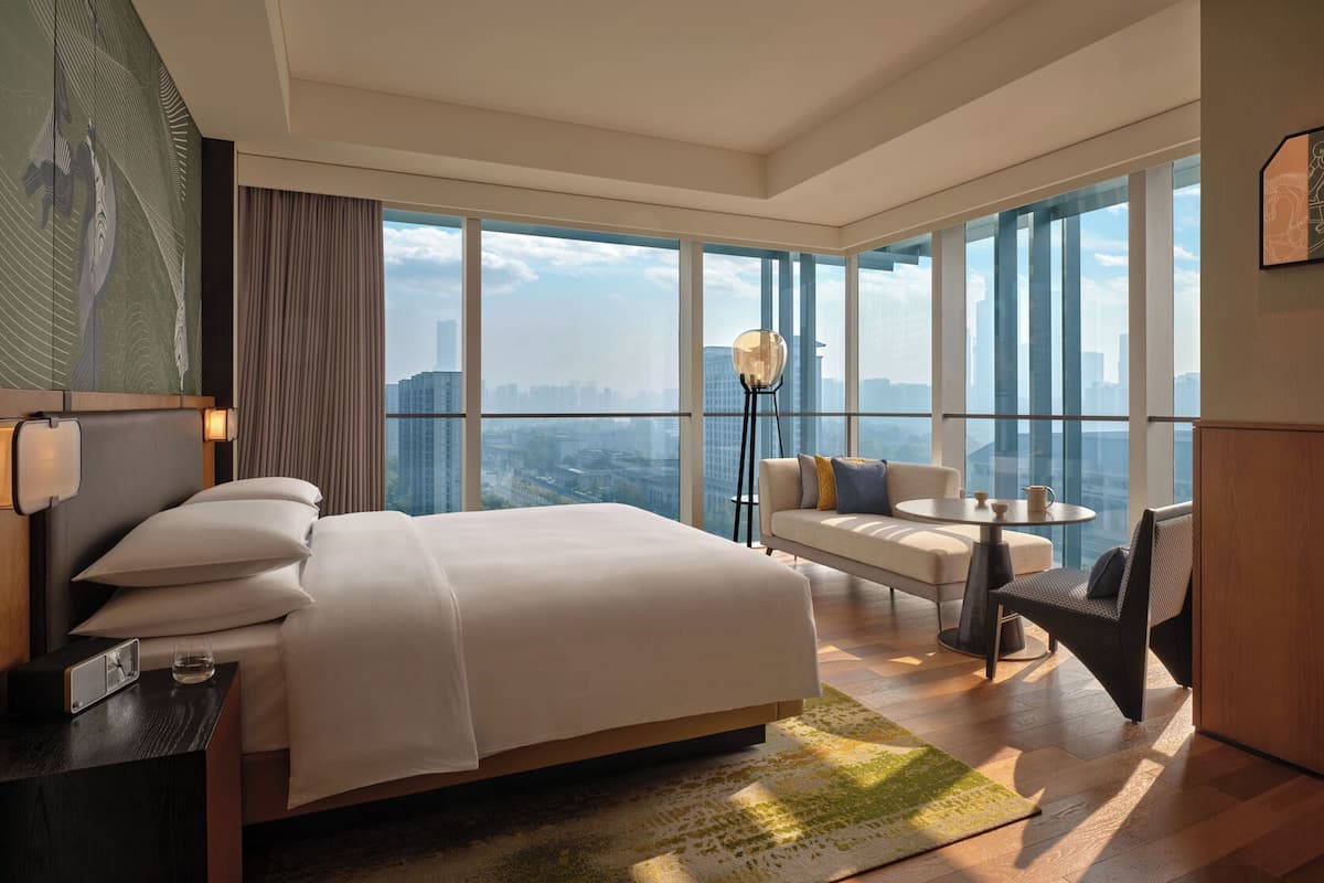 Una habitación de hotel moderna con una cama grande, un sofá, una mesita y una silla. La habitación tiene ventanas amplias que ofrecen vistas a la ciudad.