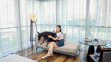 Una persona sentada en un sofá con un perro, una mesa redonda con libros y una lámpara de pie en una habitación con ventanas amplias.
