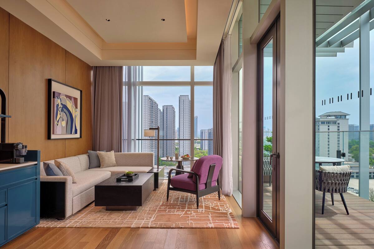 Una sala de estar moderna con sofá, mesa de centro y una silla, ventanas amplias con vista a la ciudad, y un balcón con mobiliario exterior.