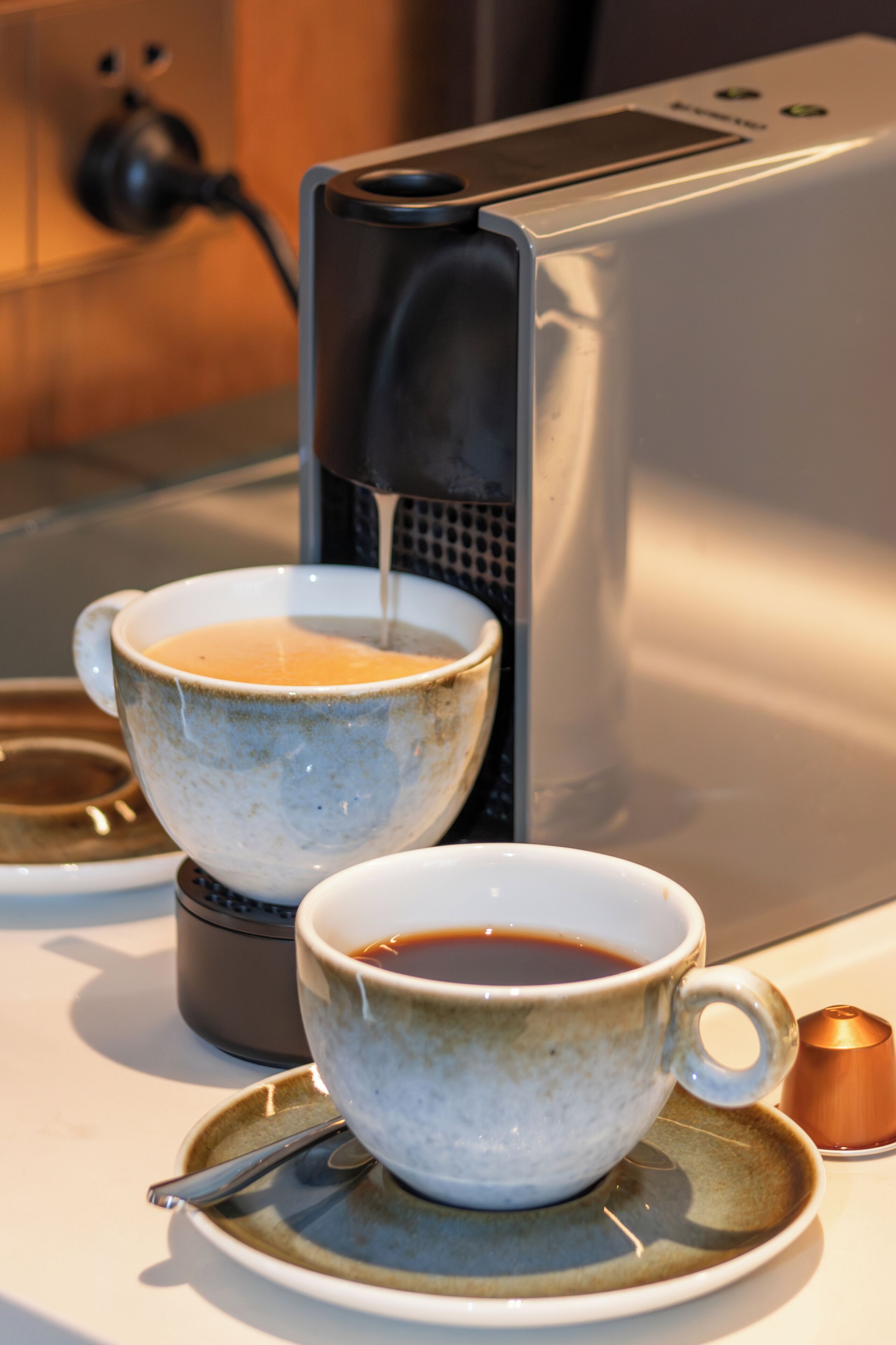 Duas chávenas de café sobre uma mesa, com uma máquina de café ao fundo.
