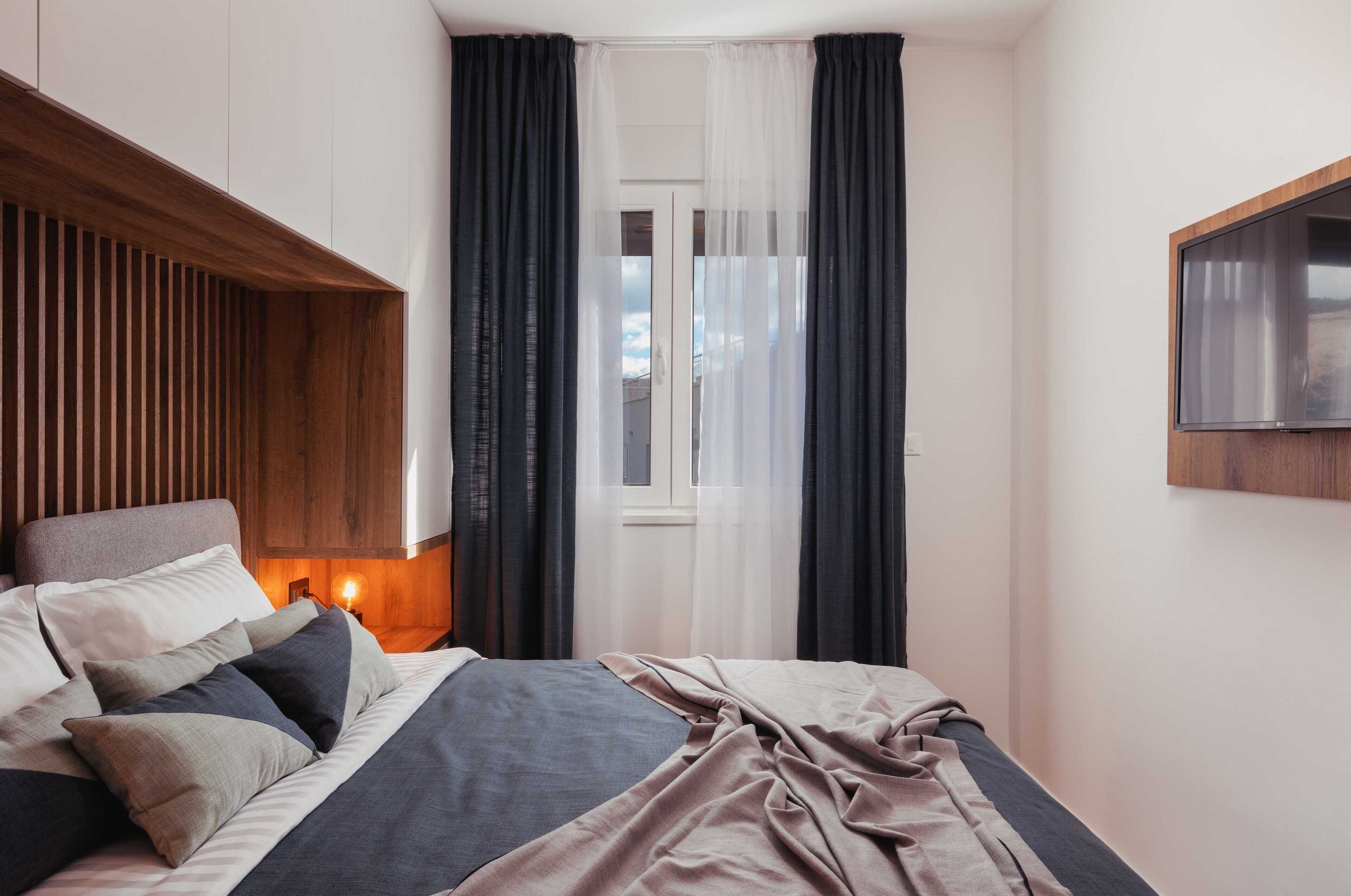 A bedroom with a bed, a nightstand with a lamp, a window with curtains, and a TV mounted on the wall.