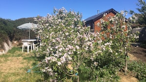 A wooden cabin with a sloped roof, surrounded by blooming trees and a clear blue sky. - Seaside Log Cabin, Carna (Carna)