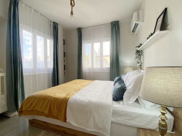 A bedroom with a bed, a bedside lamp, a window with curtains, and an air conditioning unit.
