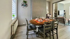 A dining area with a round wooden table, grey upholstered chairs, and a wall-mounted shelf with plants. - Daubney House – Contractors, Families, Holidays (Cleethorpes)