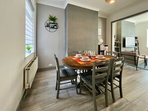 A dining area with a round wooden table, grey upholstered chairs, and a wall-mounted shelf with plants.