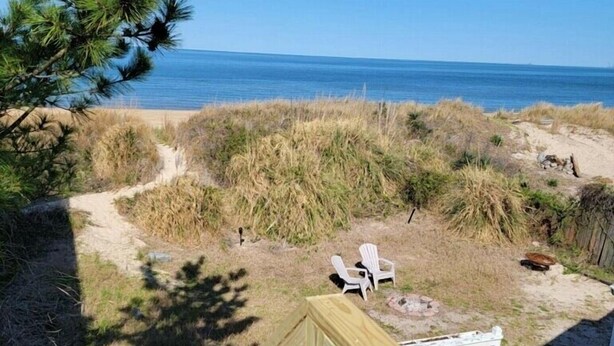 Una terraza de madera con barandilla, con vista a una playa con dunas y un cielo azul despejado.