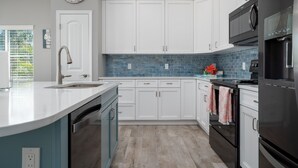 A modern kitchen with white cabinets, a blue backsplash, stainless steel appliances, and a large island with a sink. - 233 Delmar Avenue - Beautiful Private Pool Home 3 Bedroom Home by RedAwning (Fort Myers Beach)