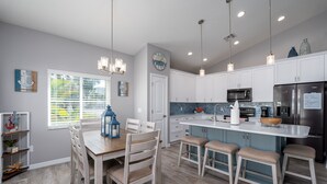 A modern kitchen with white cabinets, a central island with stools, pendant lighting, and a view of the outdoors through a window with blinds. - 233 Delmar Avenue - Beautiful Private Pool Home 3 Bedroom Home by RedAwning (Fort Myers Beach)