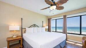 A hotel room with a large bed, a ceiling fan, a nightstand with a lamp, and a view of the beach. - Breakers 43 2 Bedroom Condo (Orange Beach)