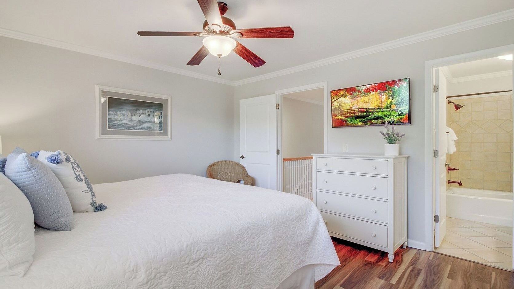 A bedroom with a bed, a ceiling fan, a dresser, a painting, and a bathroom visible through an open door.