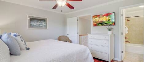 A bedroom with a bed, a ceiling fan, a dresser, a painting, and a bathroom visible through an open door.