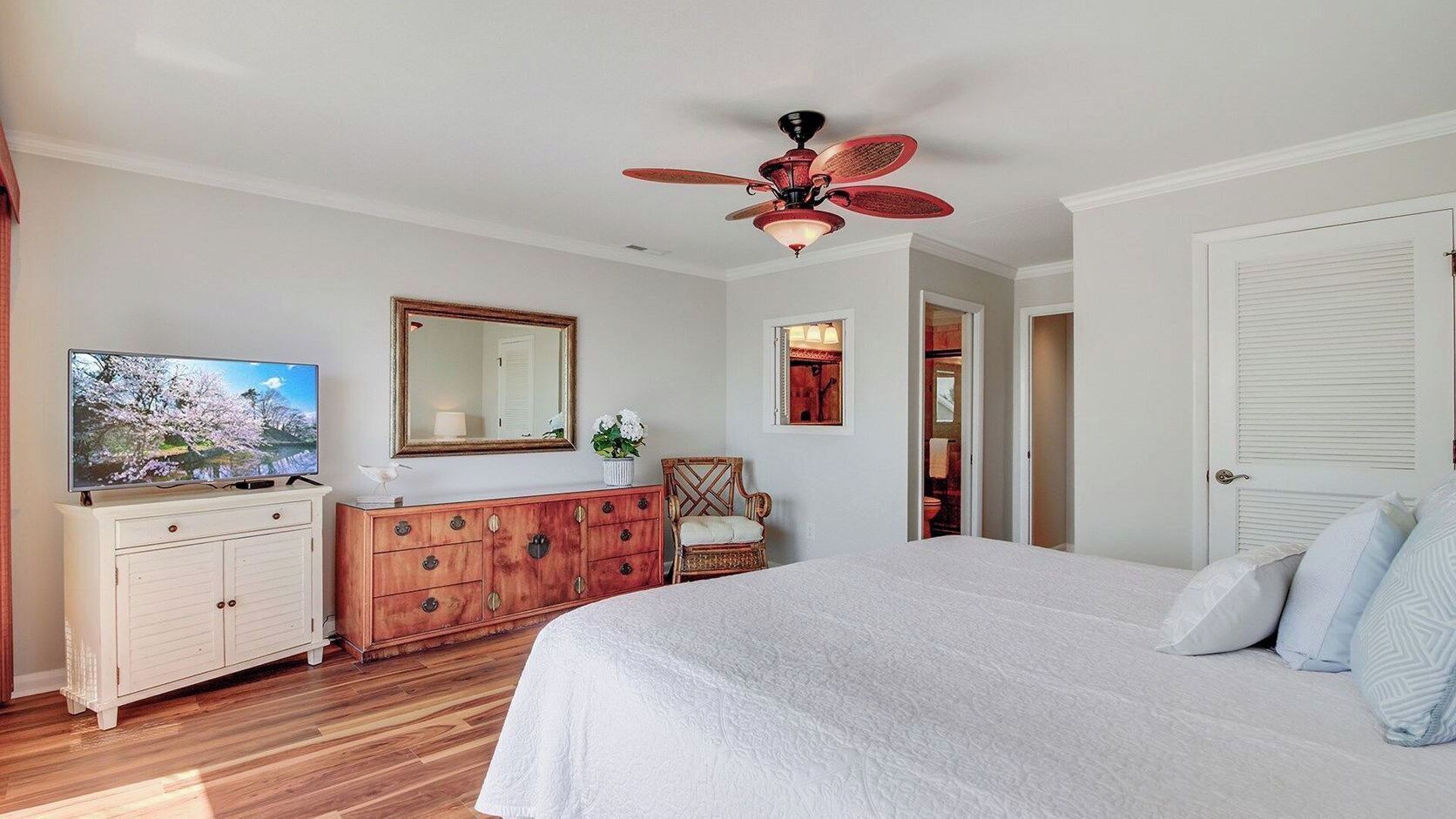 Une chambre à coucher comprenant un lit, une commode, un téléviseur, un ventilateur de plafond, un miroir et une porte.