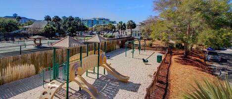 Un parque infantil con toboganes y estructuras para escalar, rodeado de arena y una valla de madera.