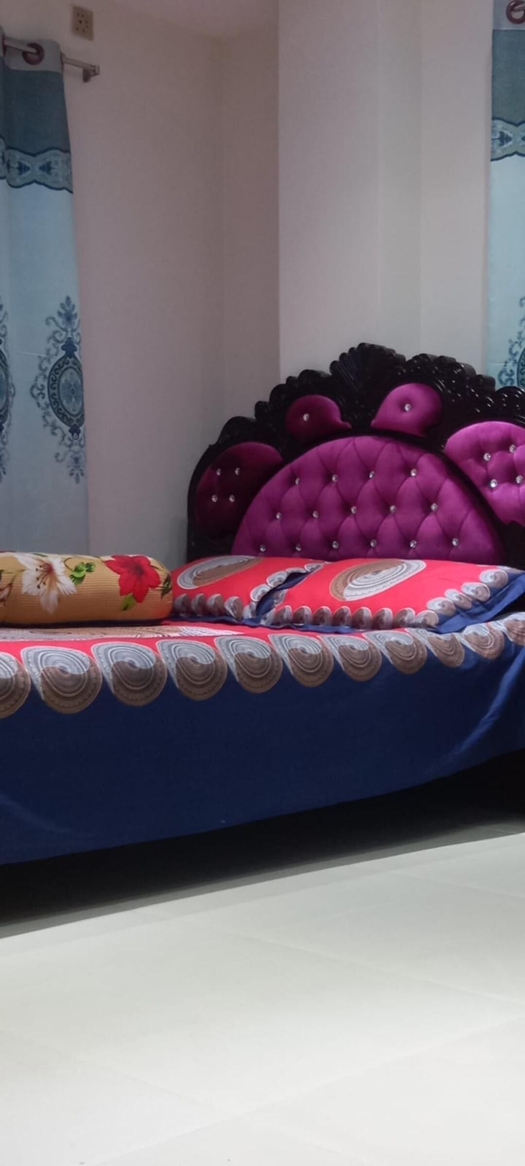 A bed with a blue bedspread and decorative pillows, a white wall, and a blue curtain with a floral pattern.