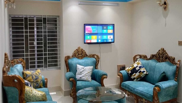 A living room with two blue armchairs, a glass coffee table, and a TV mounted on the wall. - Stunning 1-bed Apartment in Dhaka Close to Airport (Dhaka)