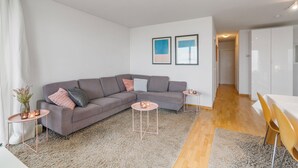 A modern living room with a grey sectional sofa, two small side tables, a wooden floor, and a white dining area with yellow chairs. - Sunrise D0 - Bright 3-bedroom apartment with large terrace and frontal sea view (De Haan)