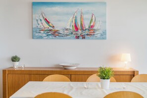 Un coin repas avec une table dressée pour quatre personnes, un tableau représentant des voiliers accroché au mur, une plante en pot et une lampe sur la table.