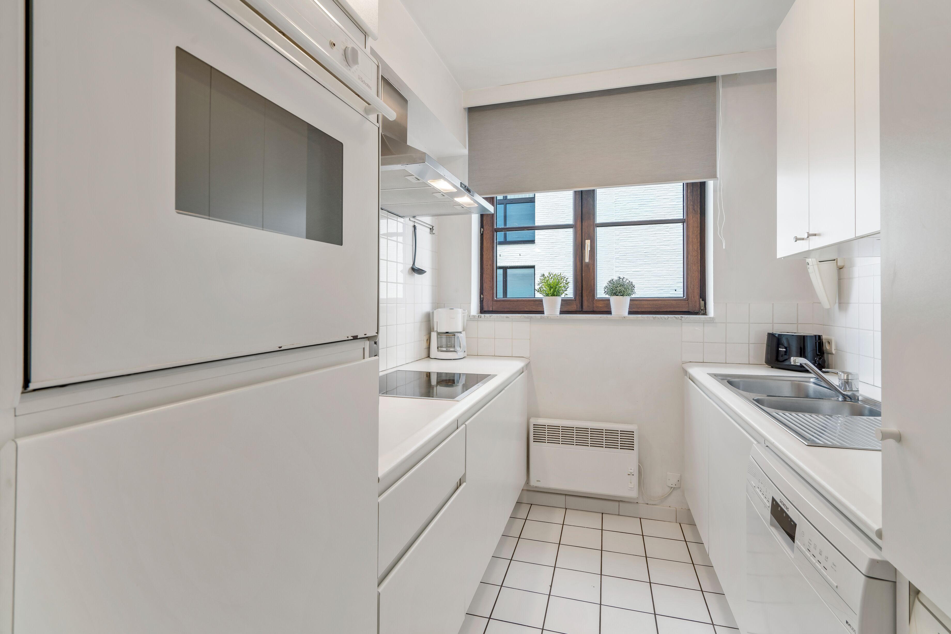 A compact kitchen with white cabinetry, a built-in oven, a coffee maker, and a window with blinds.