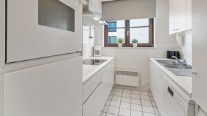 A compact kitchen with white cabinetry, a built-in oven, a coffee maker, and a window with blinds. - Sunrise D0 - Bright 3-bedroom apartment with large terrace and frontal sea view (De Haan)