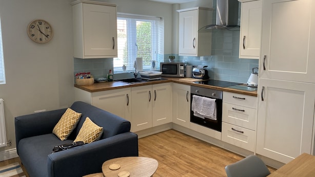 A modern kitchen with white cabinets, a microwave, a kettle, and a dining area with a blue sofa and a small wooden table. - Modern 2-bed House and Parking Tibshelf Derbyshire (Alfreton)