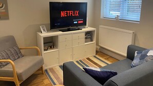 A living room with a television displaying the Netflix logo, a sofa, a chair, a radiator, and a window with blinds. - Modern 2-bed House and Parking Tibshelf Derbyshire (Alfreton)
