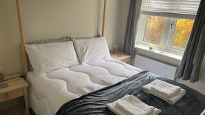 A bed with a wooden headboard, white pillows, and a black bedspread. There are two white towels on the bed. - Modern 2-bed House and Parking Tibshelf Derbyshire (Alfreton)