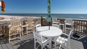 Eine Holzterrasse mit weißen Stühlen und einem Tisch, mit Blick auf einen Strand.
