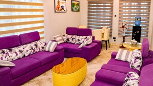 Sala de estar con sofás morados, una mesa de centro tejida de color amarillo y una silla blanca. Se observan dos cuadros en la pared y un dispensador de agua.