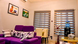 Una sala de estar con un sofá morado, una mesita y un ventilador. Hay dos ventanas con persianas estampadas y un dispensador de agua en el alféizar.