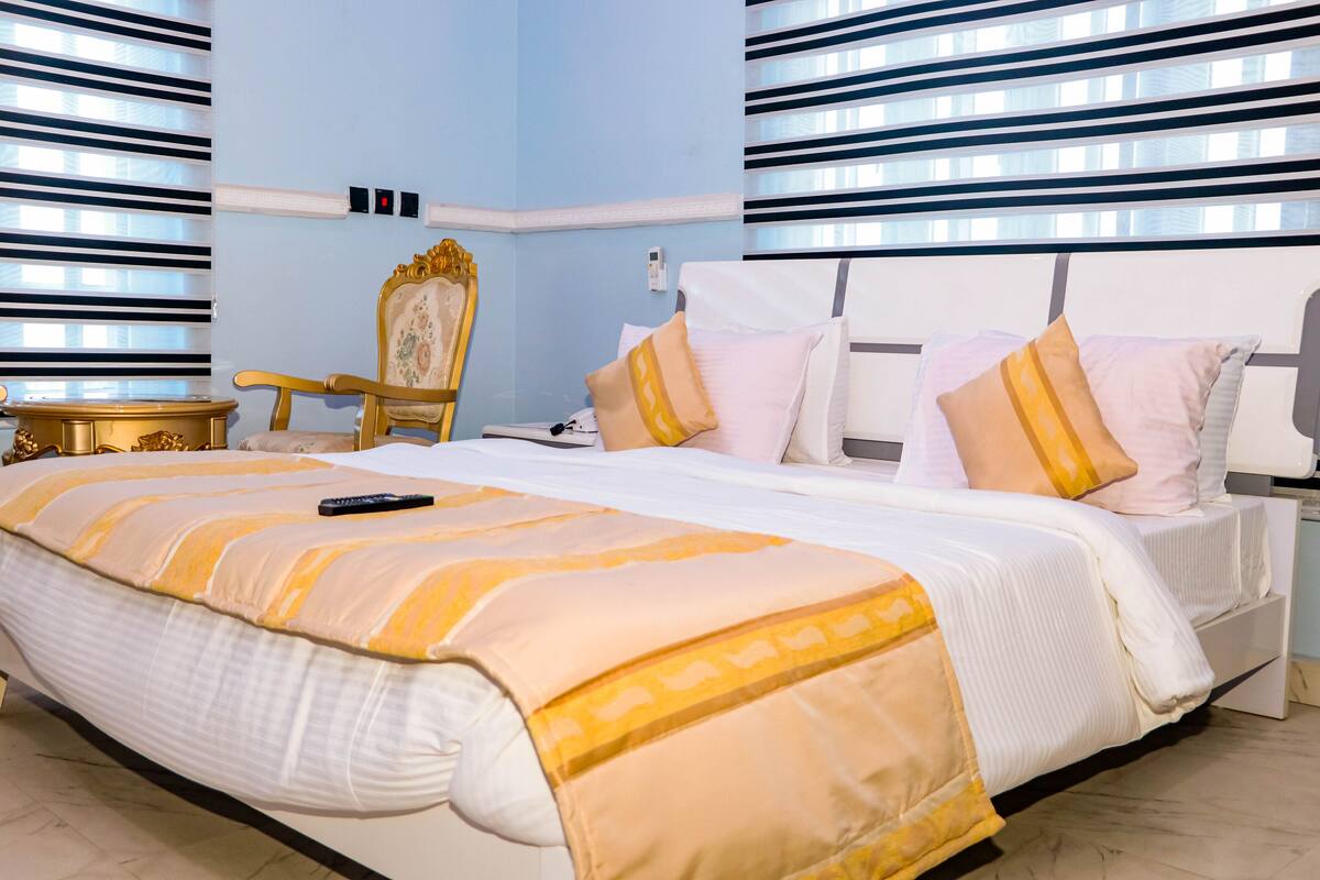 A hotel room with a large bed, a nightstand, a chair, and a window with blinds.