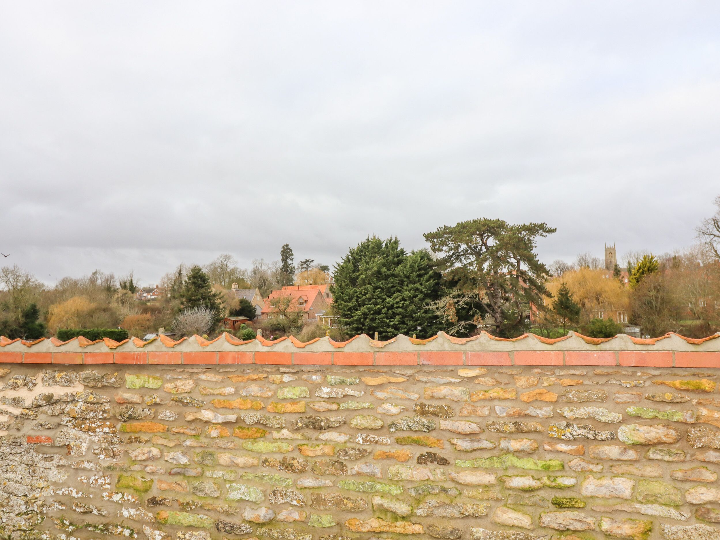 A stone wall with a tiled roof in the foreground, a tree, and houses in the background.