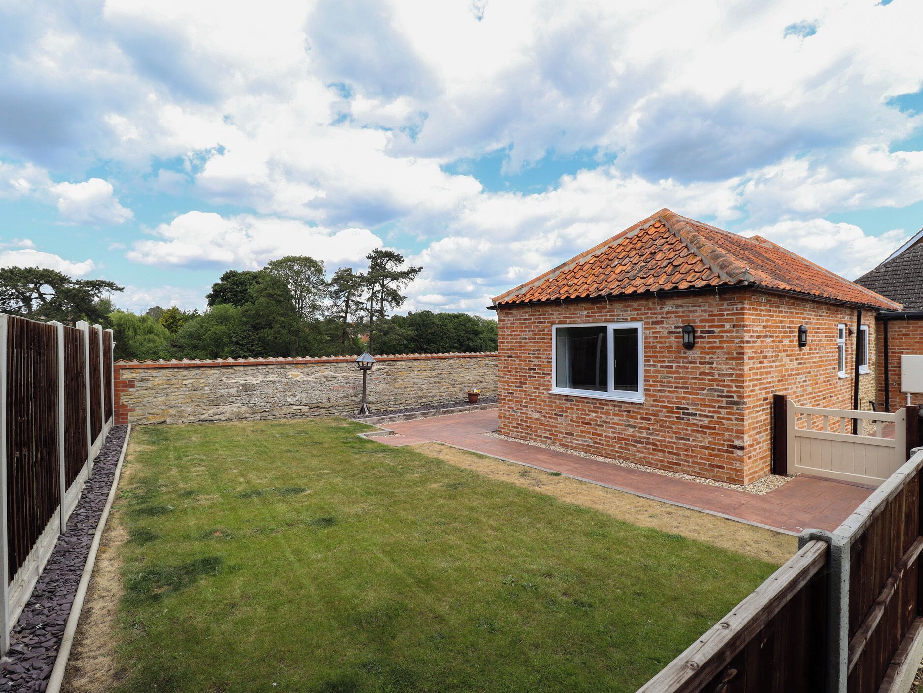 A small brick building with a tiled roof, a paved patio, and a fenced yard.