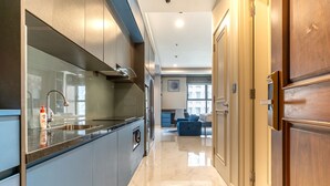 A modern kitchen with a built-in oven, a sink, and a countertop. There is a dining area with a blue sofa and a table. The floor is tiled. - The Rixx Madison (Ho Chi Minh City)