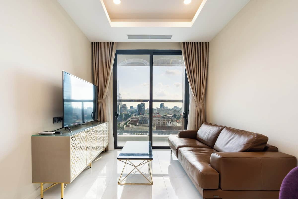 A modern living room with a leather sofa, a glass coffee table, a TV on a cabinet, and a large window with curtains.