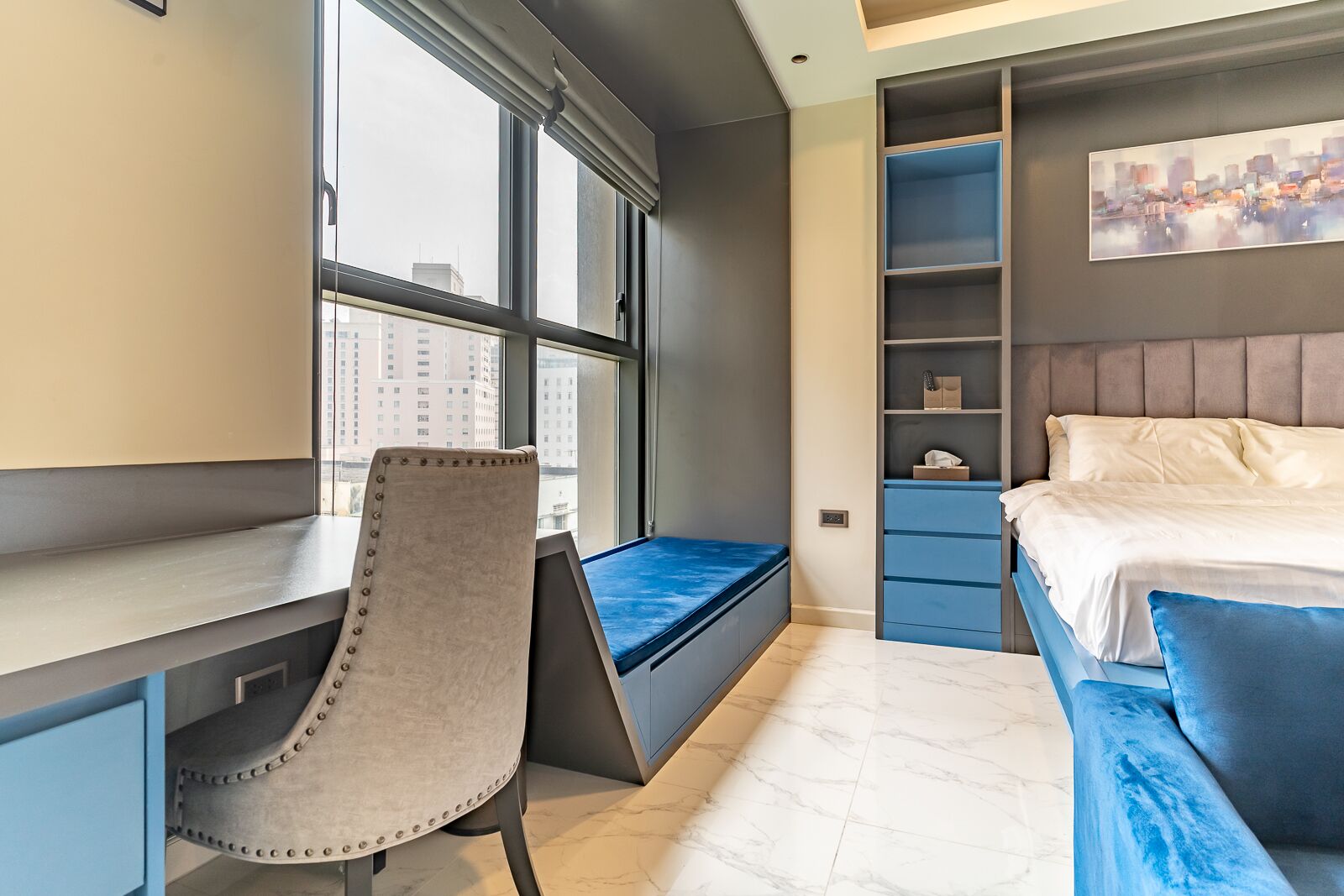 Ein modernes Schlafzimmer mit Schreibtisch, gepolstertem Sessel, Bett mit Kopfteil und Blick auf die Stadt durch das Fenster.
