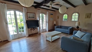 A spacious living room with a large TV, a sofa, a coffee table, and a ceiling fan. - Apartment in villa with private pool and garden, 3 bedrooms, warm welcome.  (ANTIBES)