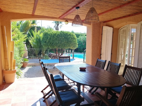 Ein Essbereich im Freien mit Blick auf einen Pool, ausgestattet mit einem runden Tisch, Stühlen, einem Reetdach und Topfpflanzen.