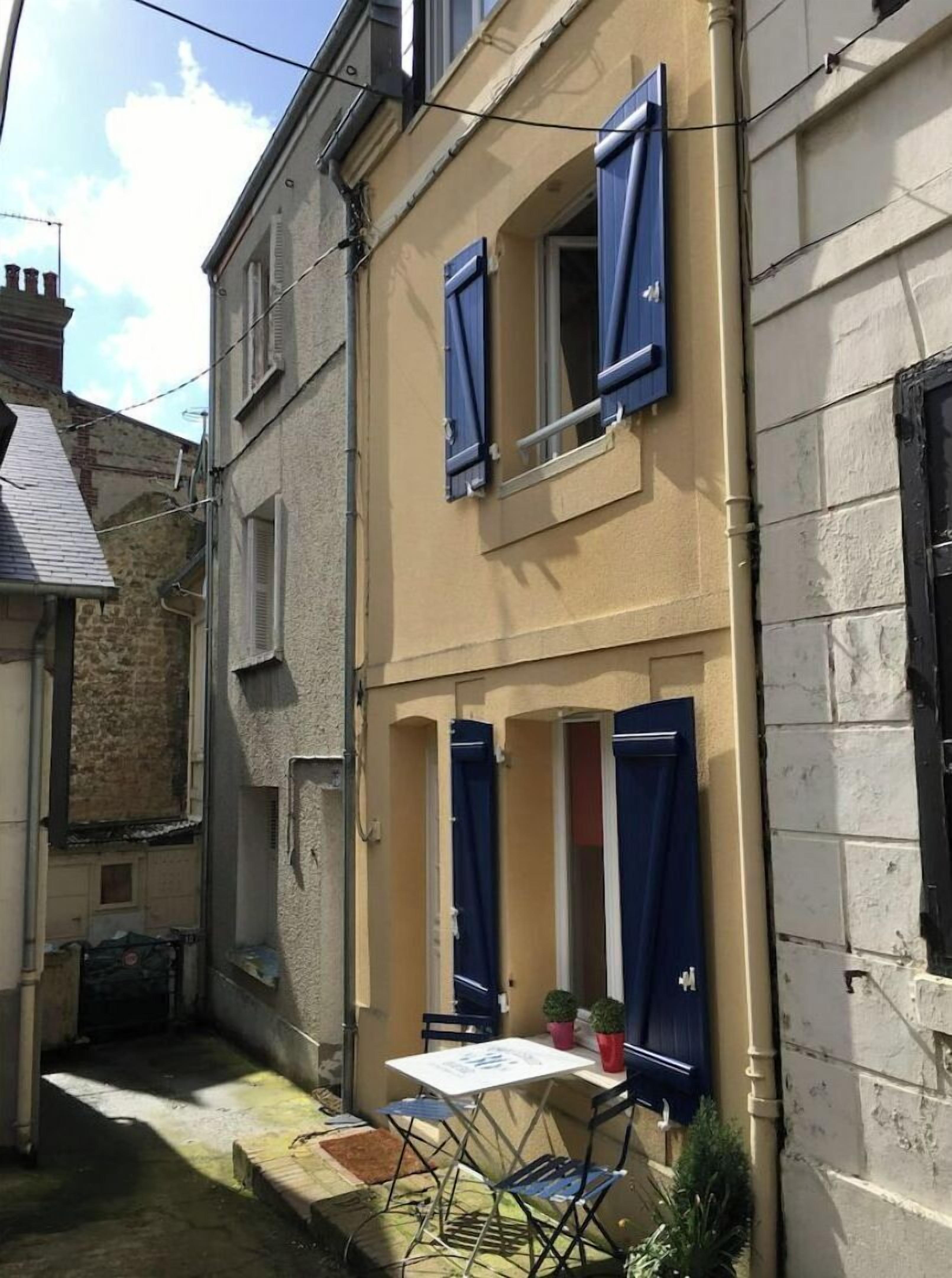 A narrow alleyway with a building featuring two windows with blue shutters, a small table with chairs, and potted plants.
