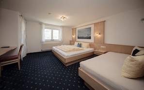 Une chambre d’hôtel avec un grand lit, un plus petit lit, un bureau et une chaise. Il y a une fenêtre avec des rideaux, un tableau au mur et un radiateur sous la fenêtre.