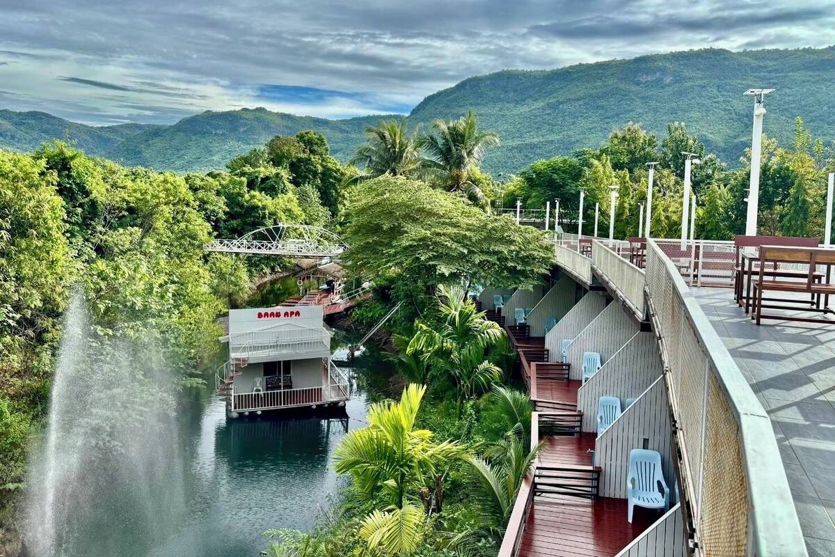 A riverside hotel with a bridge, outdoor seating, and a view of the surrounding greenery and hills.