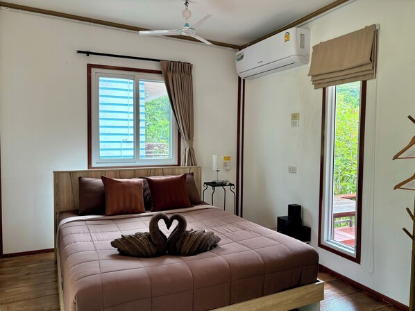 Une chambre avec un lit, un ventilateur de plafond, un appareil de climatisation, une fenêtre avec des rideaux et une vue sur la verdure à l’extérieur.