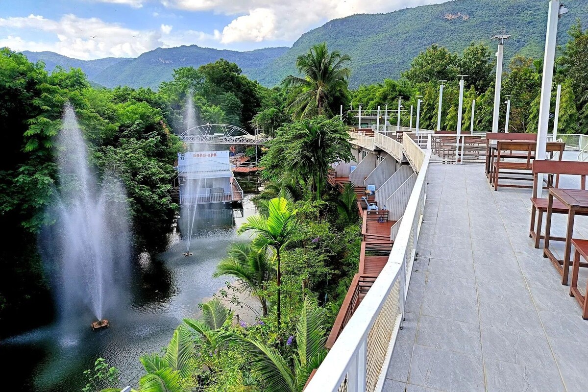 Un paseo a orillas del río con un puente, una cascada y una zona de comedor con mesas y sillas.