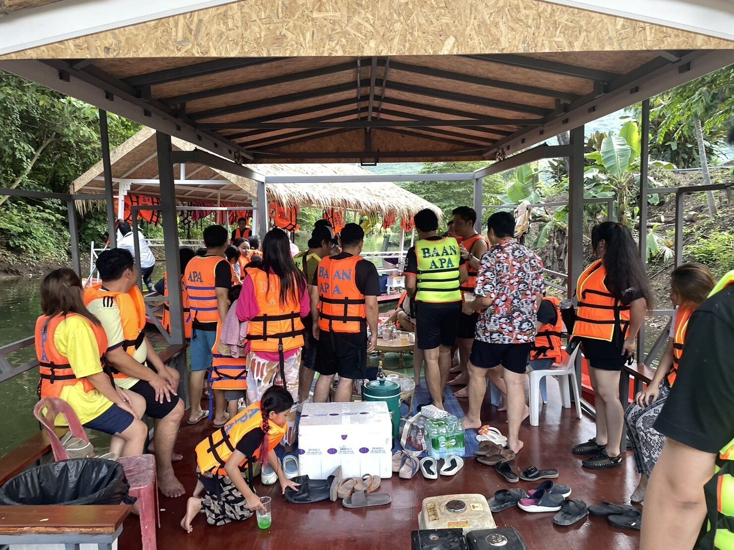 Un grupo de personas con chalecos salvavidas en un bote, algunas de pie y otras sentadas, con varios objetos como zapatos y cajas en el suelo.