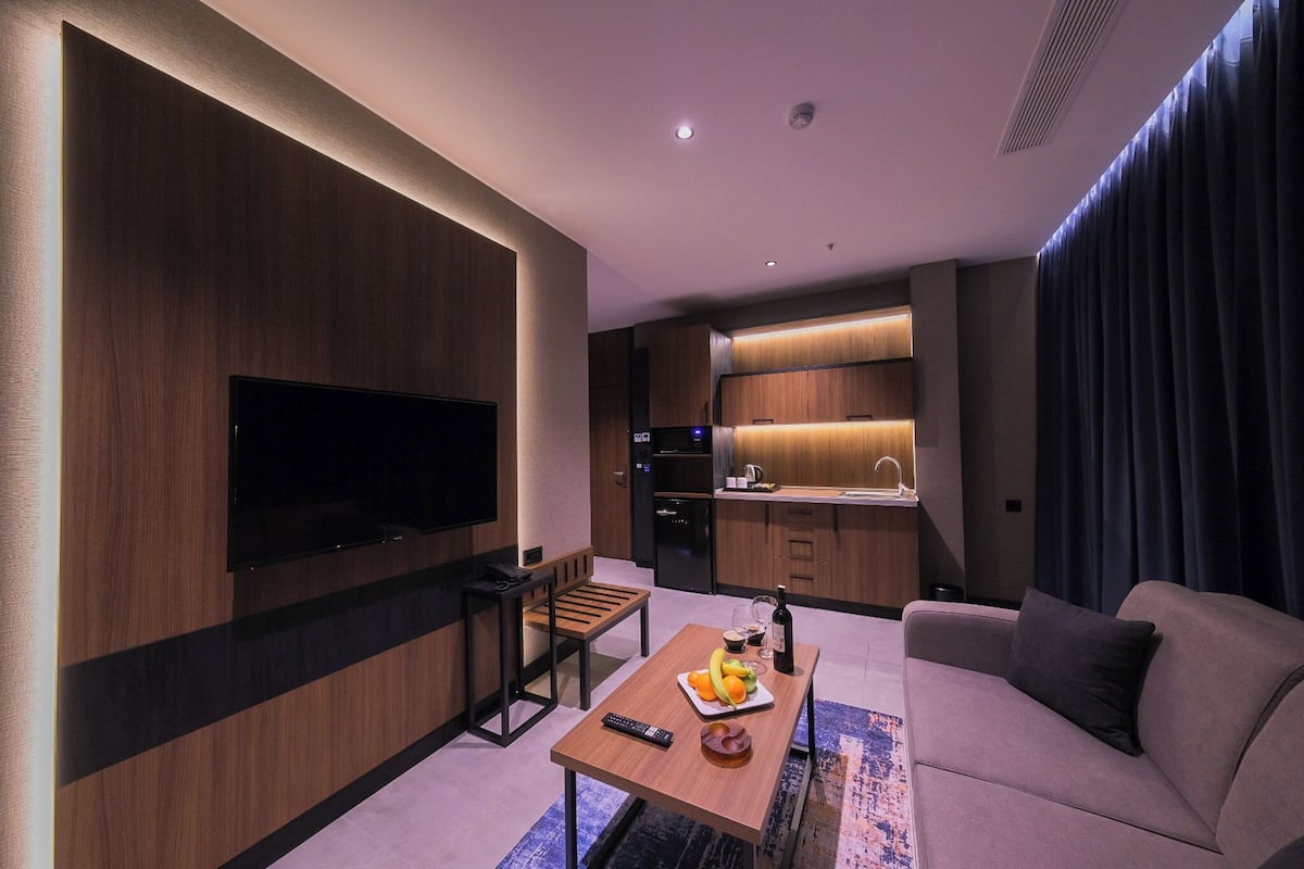 Una sala de estar moderna con un televisor de pantalla plana, un sofá, una mesa de centro con frutas y una botella, un banco de madera, y un área de cocina con un lavabo y armarios.