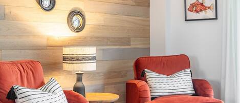 A modern living room with two red velvet armchairs, a round wooden coffee table, and a floor lamp. The wall features wooden panels and framed artwork.