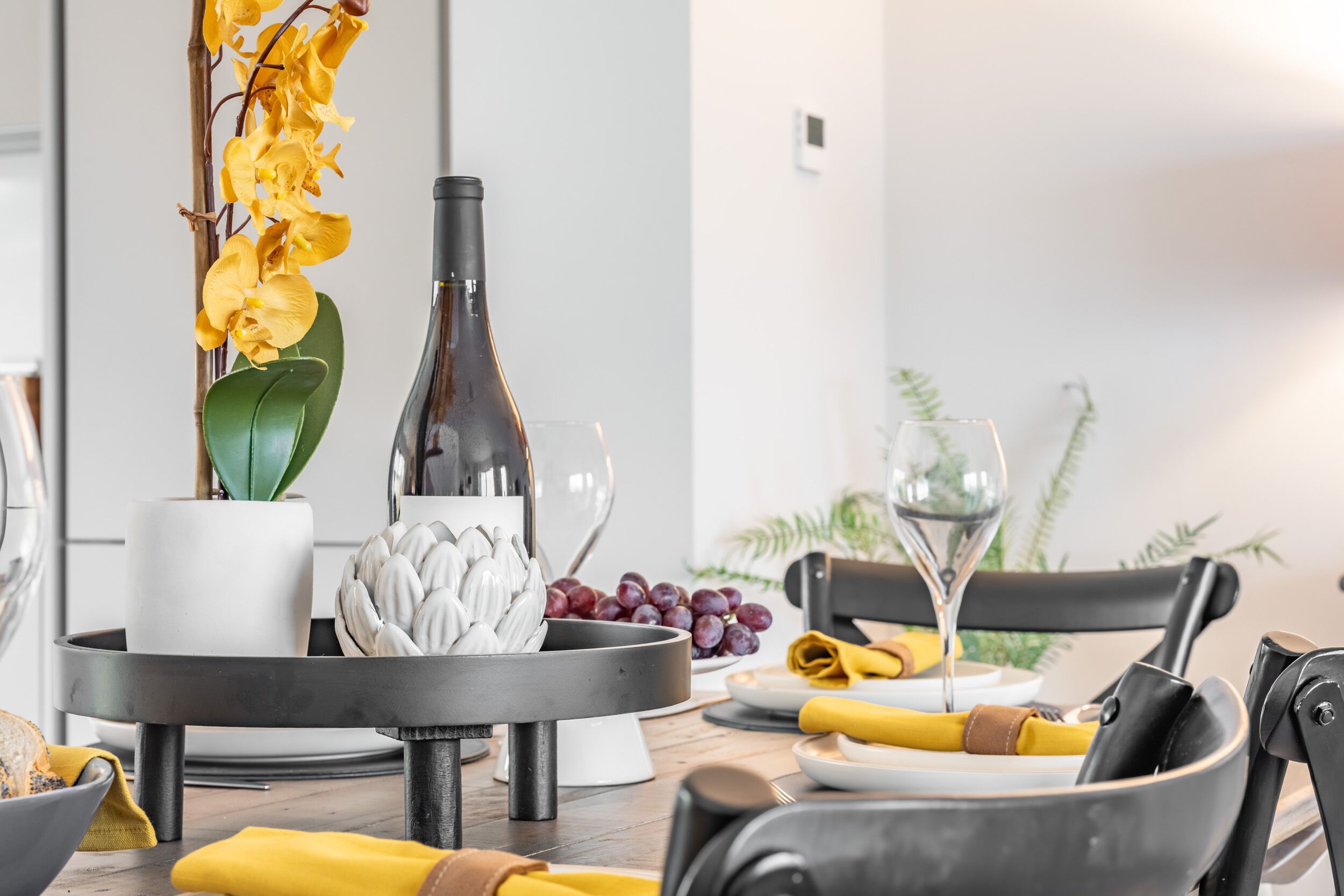 A dining table with a bottle, a bowl of grapes, and a vase of yellow flowers.