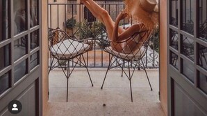 Varanda com uma pessoa relaxando, uma palmeira e um edifício com telhado de azulejos ao fundo.
