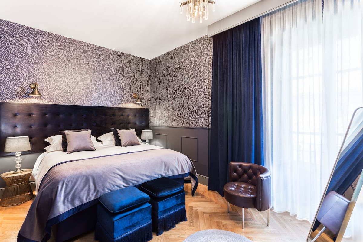 Un dormitorio moderno con una cama grande, un cabecero capitonado, una mesita de noche, un sillón de cuero y un amplio ventanal con cortinas.