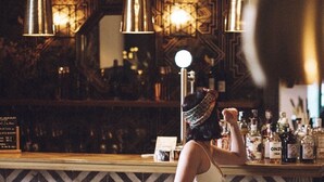 Uma área de bar com uma mulher sentada no balcão, uma parede estampada e um candelabro.