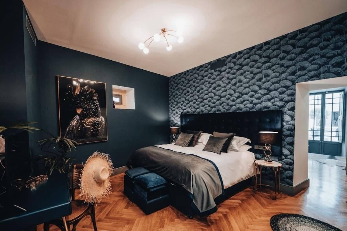 Un dormitorio moderno con una pared azul oscuro, una cama grande con muchas almohadas, una mesita de noche pequeña y una lámpara colgante.